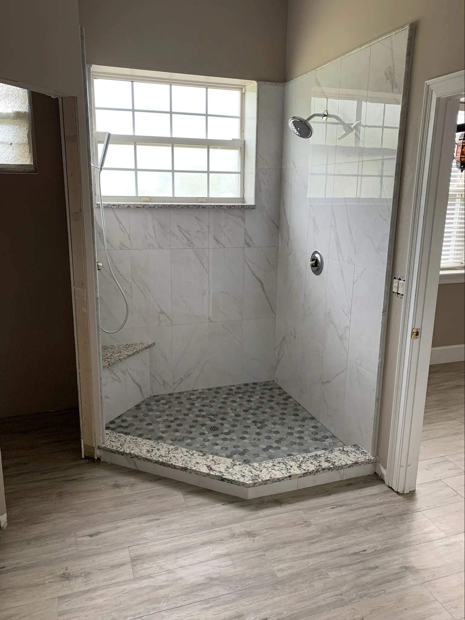 Compact walk-in shower with marble-look wall tile, pebble mosaic floor, window, and stone curb.