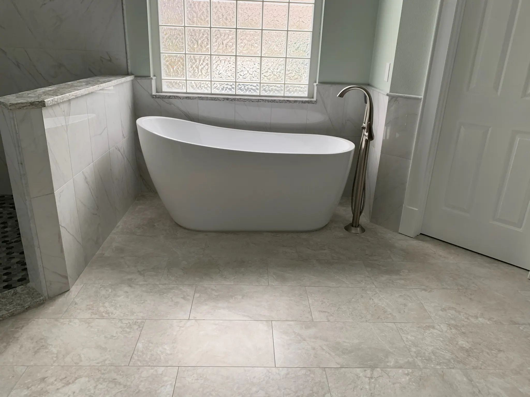 Freestanding white tub beside a tiled half-wall and walk-in shower with pebble flooring.