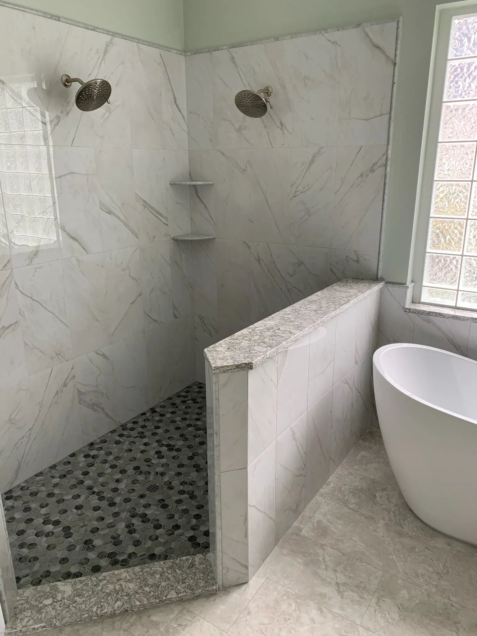 Marble-look walk-in shower with corner shelves, pebble floor, and half-wall beside a tub.