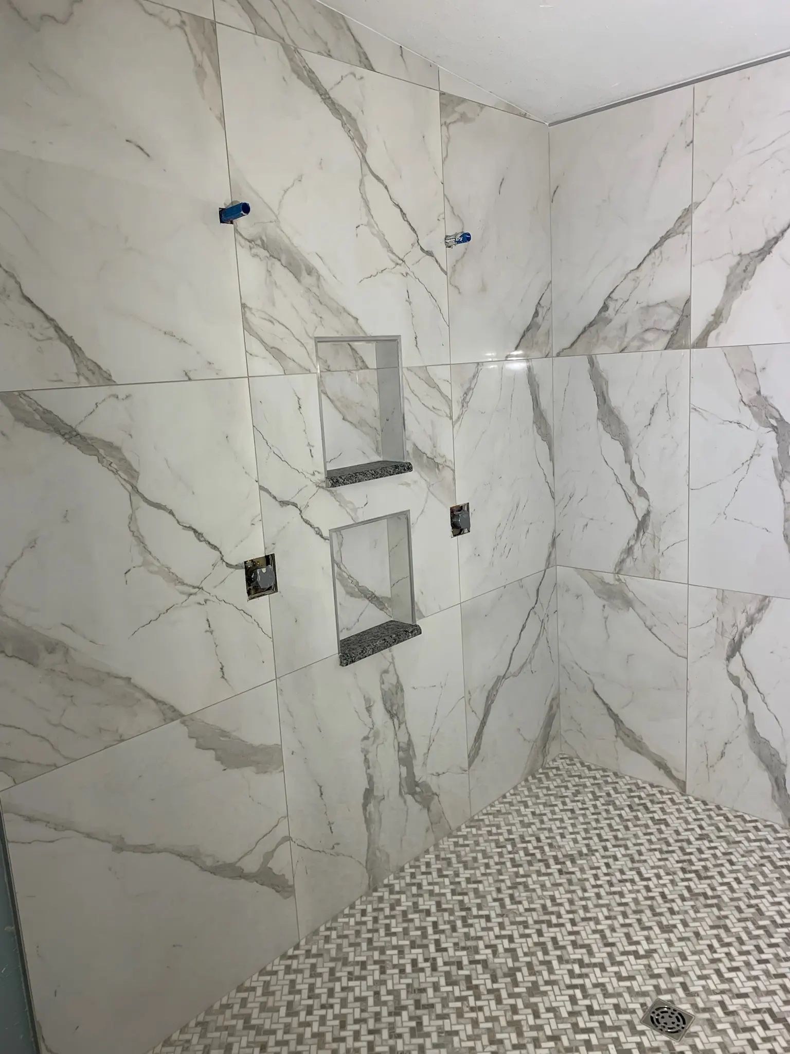 Large-format marble-look shower walls with two recessed niches and basketweave mosaic flooring.