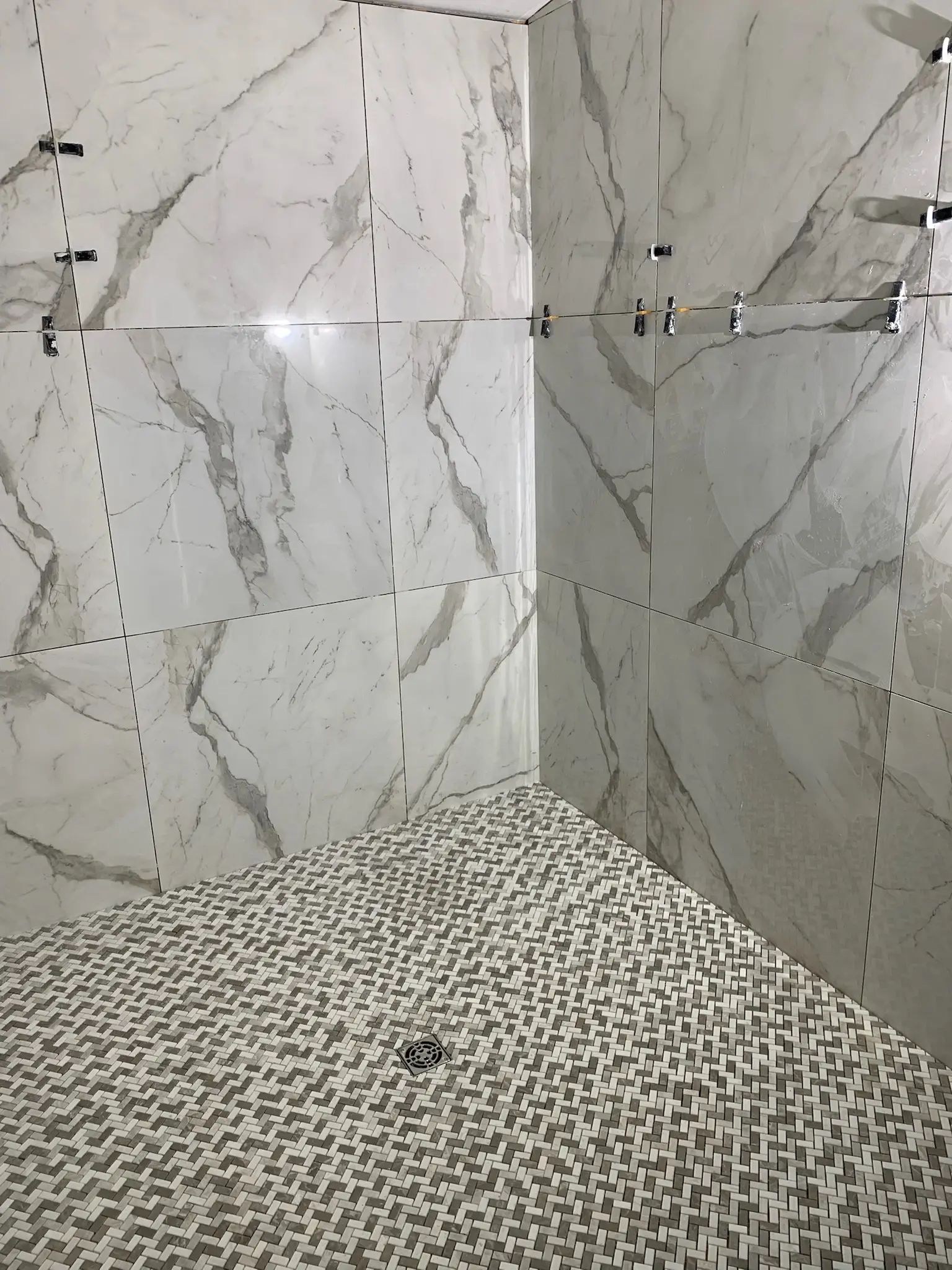Corner view of a marble-look shower with basketweave floor tile and floating shelf.