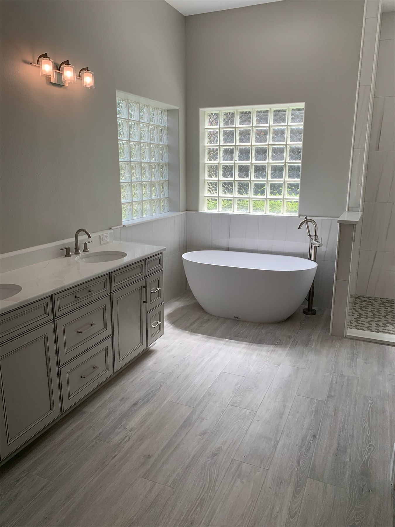Gray double vanity and freestanding tub in a bright bathroom with wood-look floor tile.