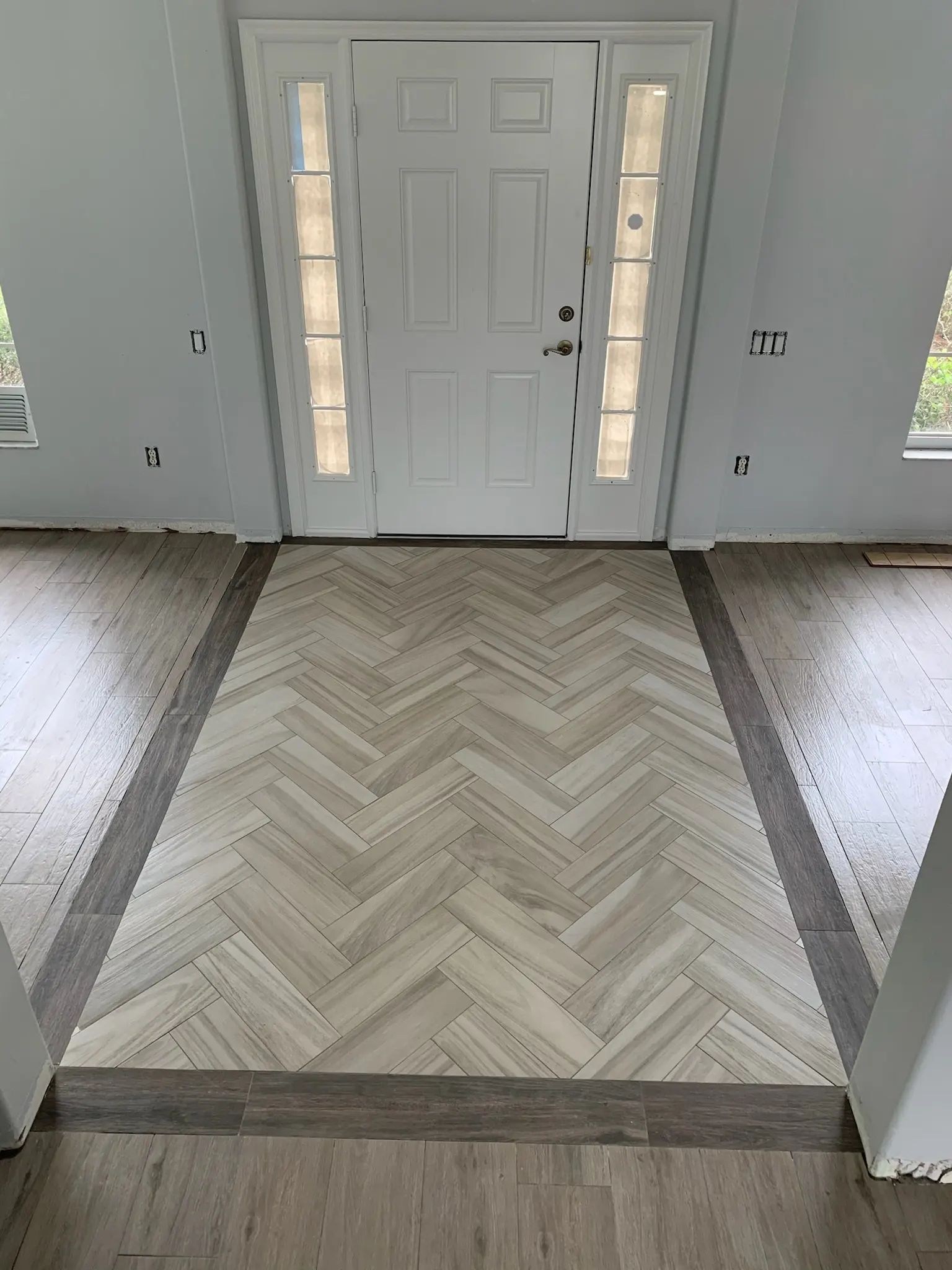 Front entry with a light herringbone tile inset, dark framed border, and surrounding wood-look planks.