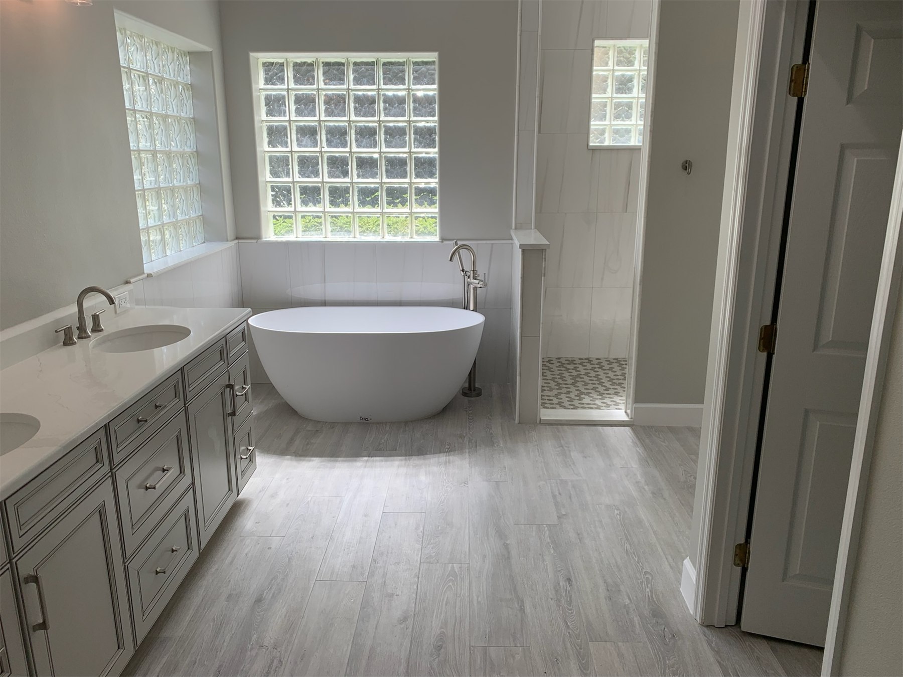 Wide view of a light-filled bathroom with a gray double vanity, freestanding tub, and tiled shower.