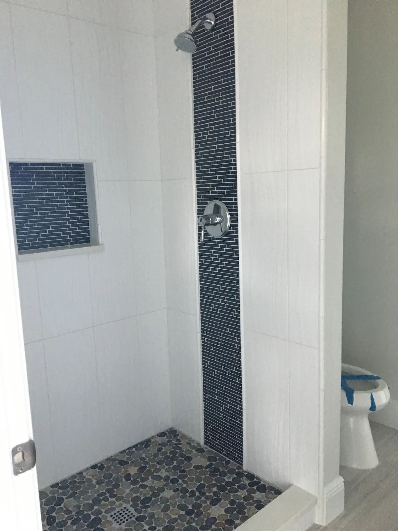 White shower with a blue glass mosaic stripe, matching framed niche, and multitone pebble floor.