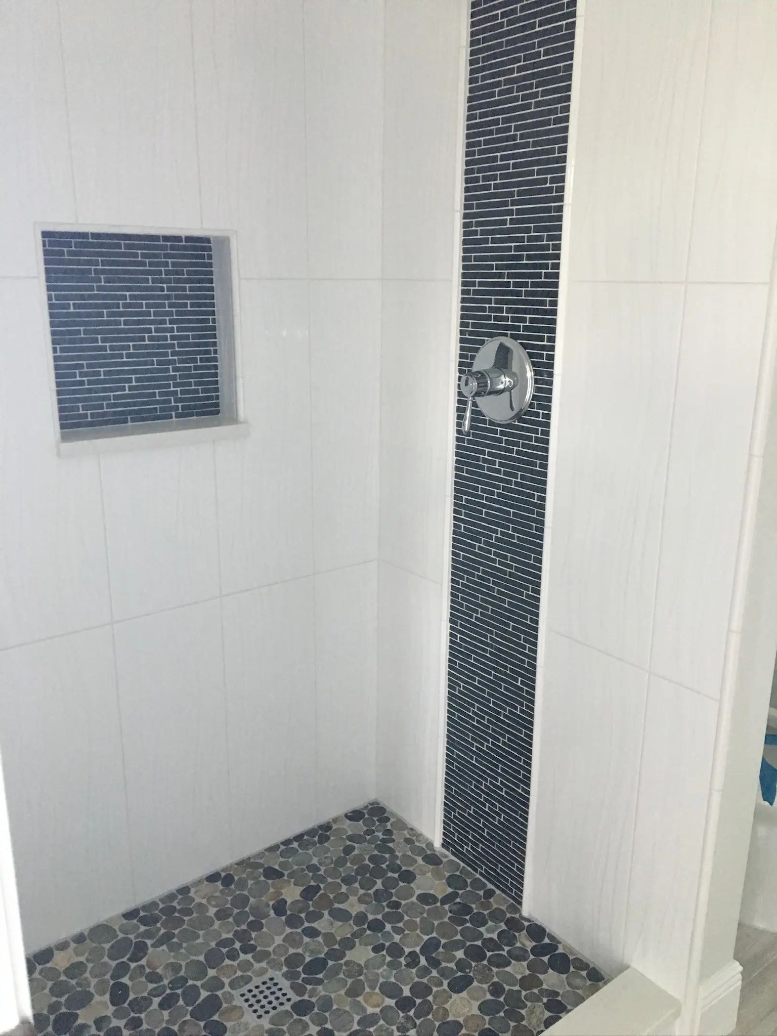 Shower entry view showing white wall tile, blue glass mosaic details, and pebble flooring.