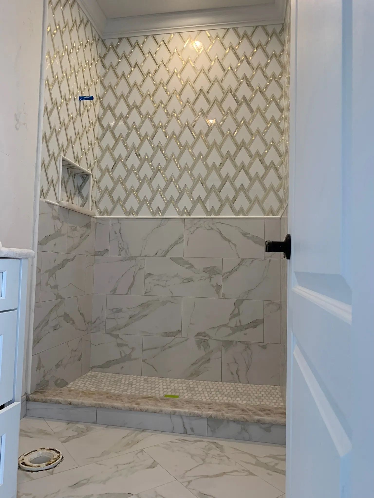 Angled view through a shower doorway showing geometric mosaic tile, marble-look wall tile, and pebble flooring.