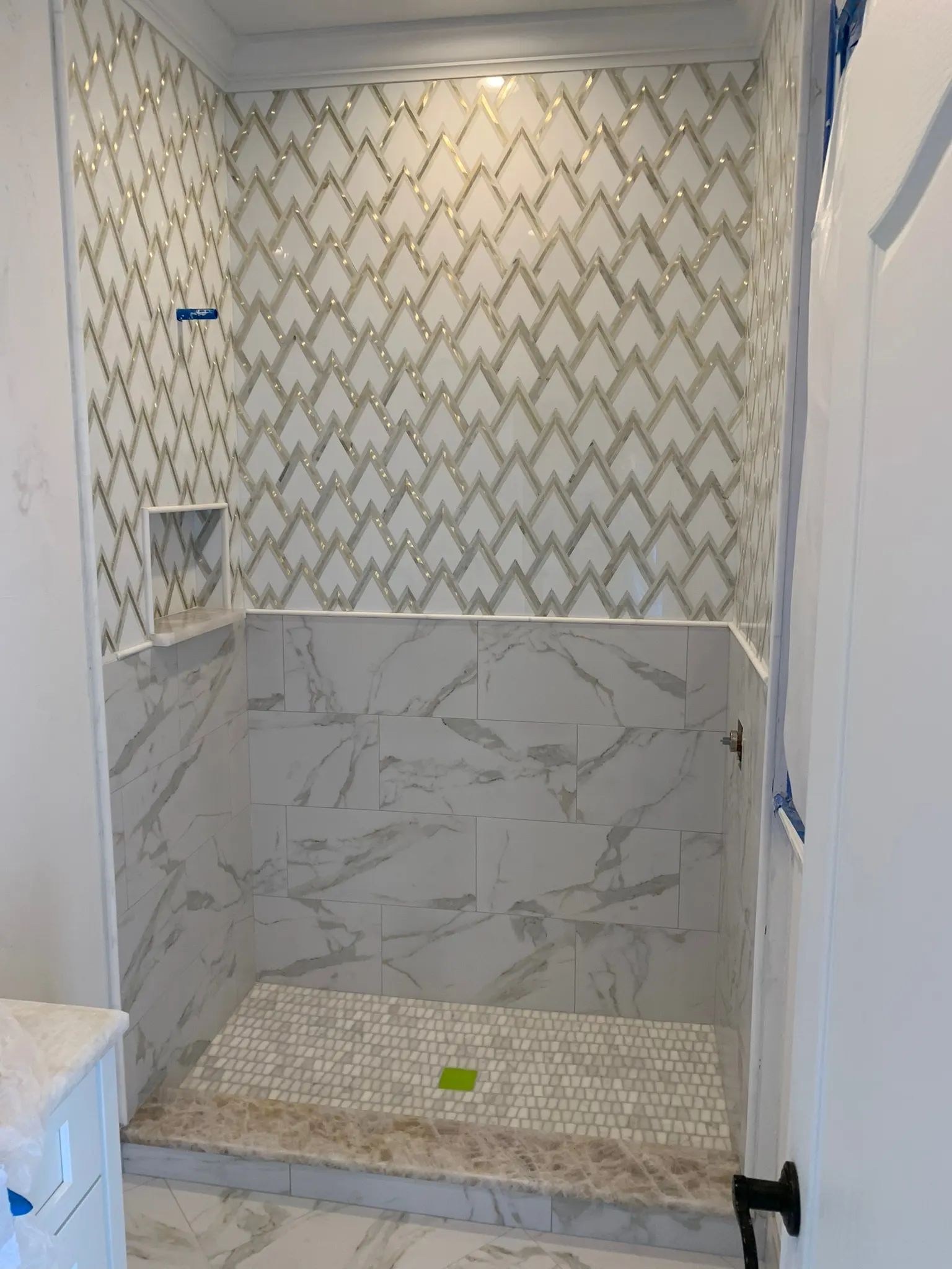 Walk-in shower with gray geometric mosaic walls, marble-look lower tile, and a pebble mosaic floor.